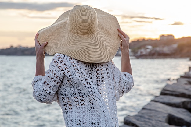 Фото Красивая молодая женщина у моря на закате. одета в длинную юбку и шляпу.
