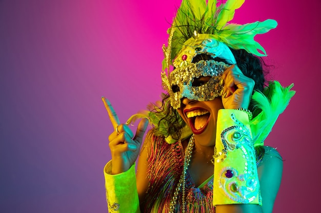 Beautiful young woman in carnival and masquerade costume on gradient studio background in neon light