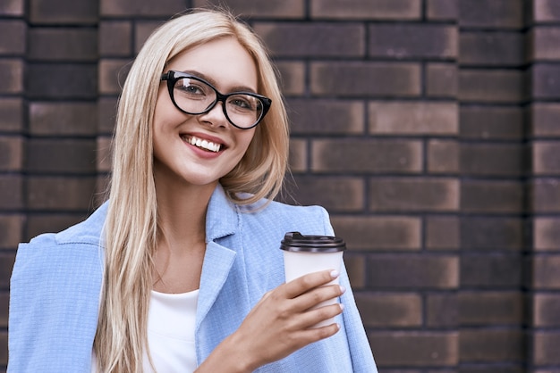 La bella giovane donna in abiti casual e occhiali tiene in mano una tazza di caffè e