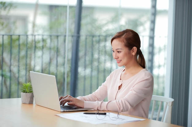 Beautiful young woman in casual clothing using laptop and smiling while working indoors