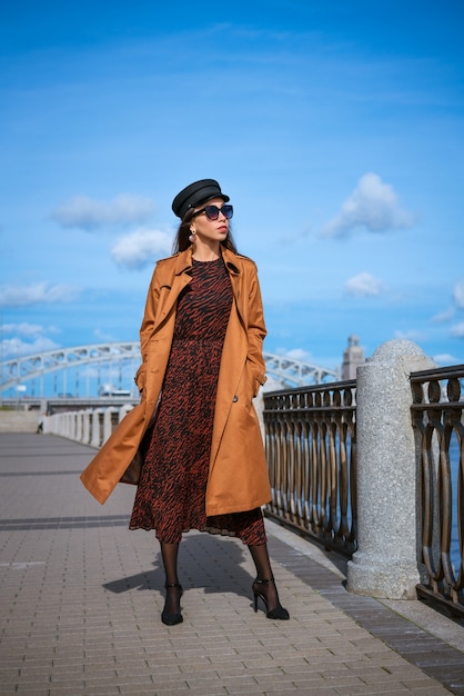 Beautiful young woman of caucasian appearance in a brown raincoat and dress posing on the embankment...