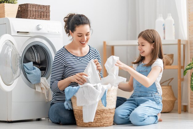 美しい若い女性と子供の女の子の小さなヘルパーは、家で洗濯をしながら楽しんで笑っています。