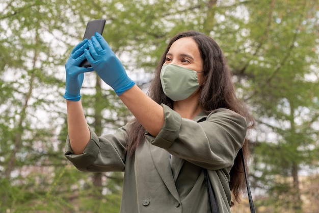 Красивая молодая женщина в тканевой маске и латексных перчатках с помощью смартфона, принимая селфи в маске на открытом воздухе