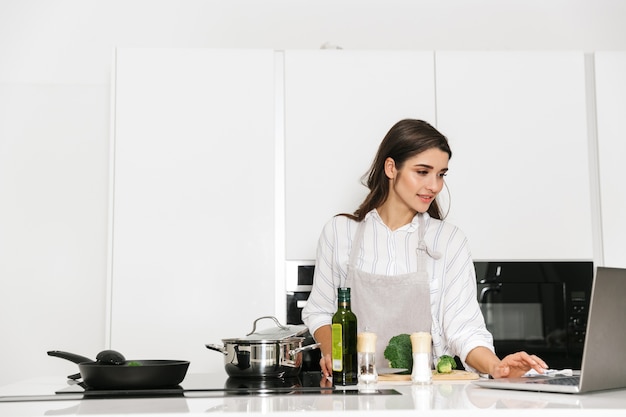 ラップトップコンピューターを見て、キッチンで鍋で健康的な夕食を調理する美しい若い女性