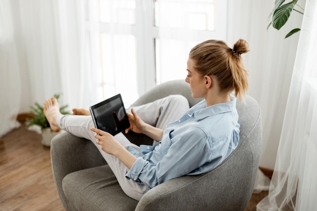 beautiful young woman in cozy home clothes is working on a tablet at home in the living room
