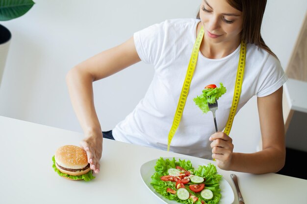 Photo beautiful young woman decides eating hamburger or fresh salad in kitchen