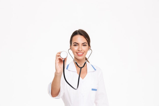 Beautiful young woman doctor wearing uniform standing isolated over white wall, using stethoscope