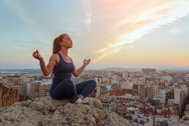 Beautiful young woman does yoga outdoors, on top of a mountain ridge in the city.