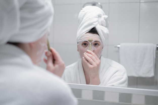 Bella giovane donna che fa massaggio facciale con lo strumento gua sha davanti allo specchio a casa