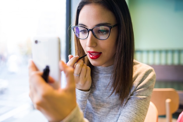 Beautiful young woman doing make-up with her lipstick and mobile phone.