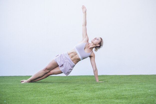 庭の芝生でストレッチ運動をしている美しい若い女性。運動をしながらバランスをとるスポーツウェアの女性。緑の芝生の上で屋外でヨガを練習するスポーティな若い女性