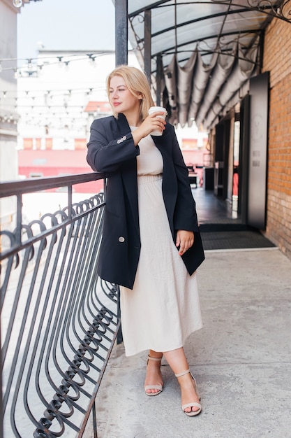 Beautiful young woman in dress and jacket walks around the city and drinks coffee