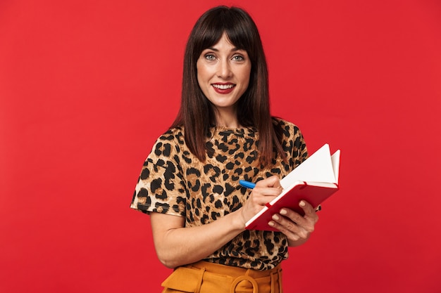 beautiful young woman dressed in animal printed shirt posing isolated over red wall writing notes in notebook.