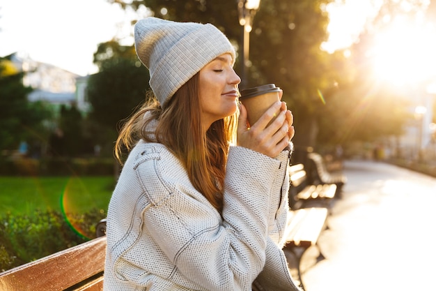 Beautiful young woman dressed in autumn coat and hat sitting on a bench outdoors, holding cup of warm coffee