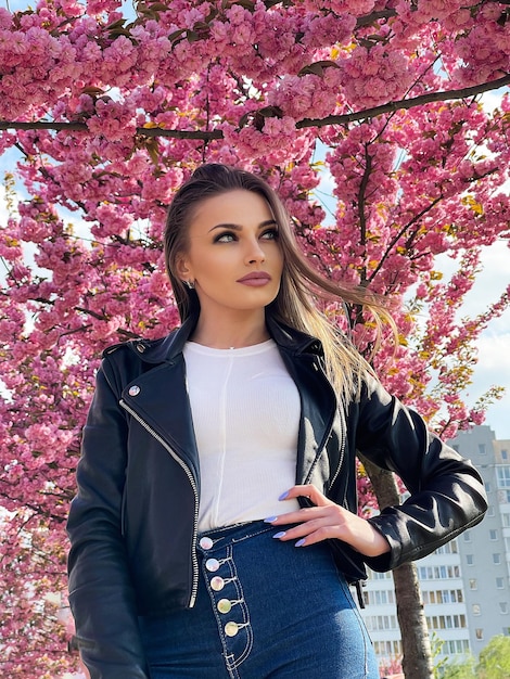 Beautiful young woman dressed in black jacket stands in the color of sakura flowers in the spring