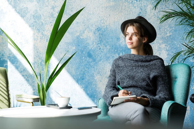 Beautiful young woman dressed in sweater and hat sitting in chair at the cafe table, taking notes