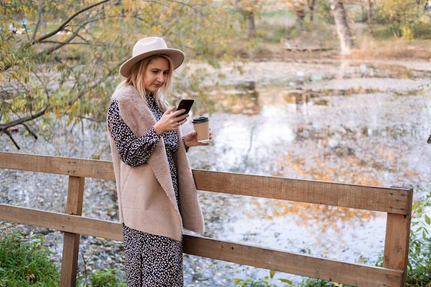 Bella giovane donna che beve caffè, parlando al telefono in natura in riva al lago nel parco autunnale in autunno