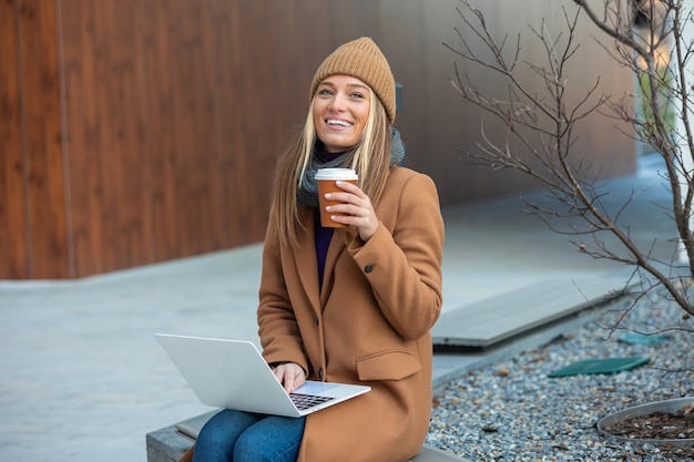 Beautiful young woman drinking cup of coffee outside on street cafe working on laptop