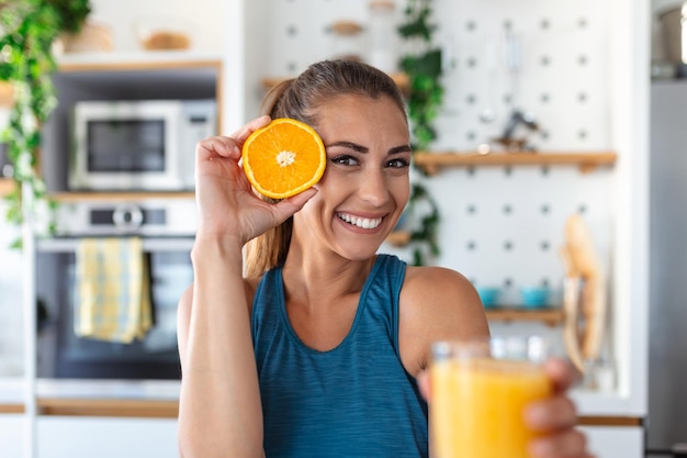 Beautiful young woman drinking fresh orange juice in kitchen Healthy diet Happy young woman with glass of juice and orange at table in kitchen