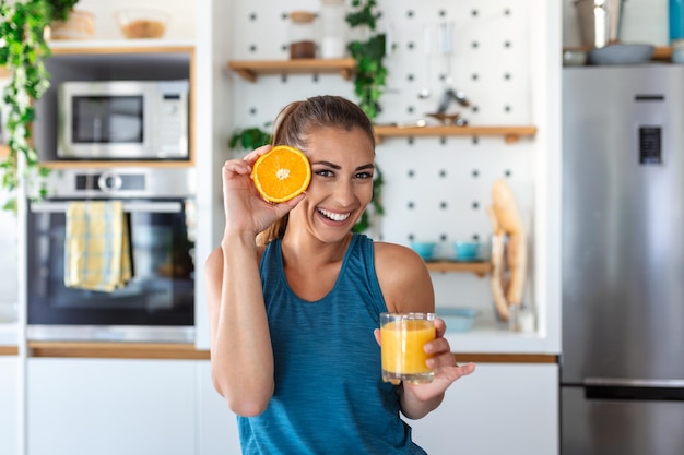 Beautiful young woman drinking fresh orange juice in kitchen Healthy diet Happy young woman with glass of juice and orange at table in kitchen