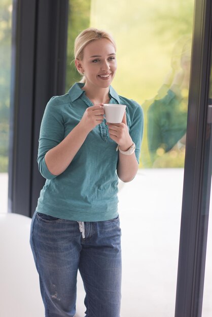 彼女の家の窓のそばで朝のコーヒーを飲む美しい若い女性