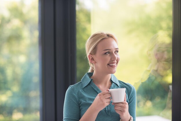 彼女の家の窓のそばで朝のコーヒーを飲む美しい若い女性