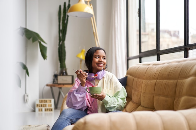 Beautiful young woman eating cereal in a bowl sitting on the sofa at home.