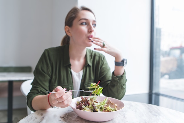 Beautiful young woman eating salad in restaurant and looking in window. Focus on salad plate.