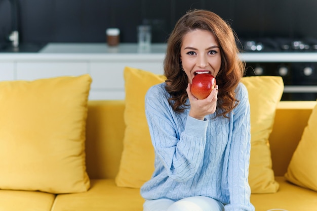 Beautiful young woman eats juicy red apple in the modern cuisine and posing on camera with pleasant