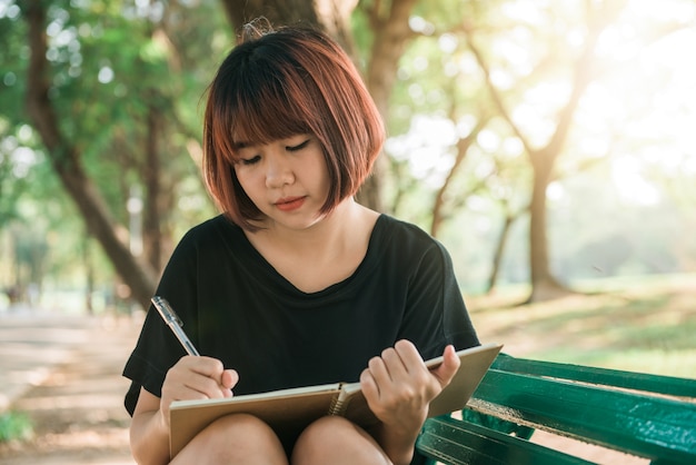 ノートブックで何かを書く庭のベンチで夏を楽しむ美しい若い女性