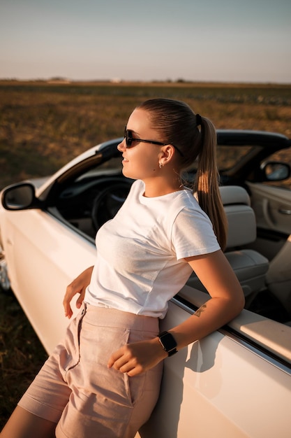 선글라스의 얼굴에 유럽풍의 아름다운 젊은 여성, 그녀는 흰색 컨버터블 근처에 서 있습니다. 성공적인 여성 사업가와 그녀의 흰색 차.