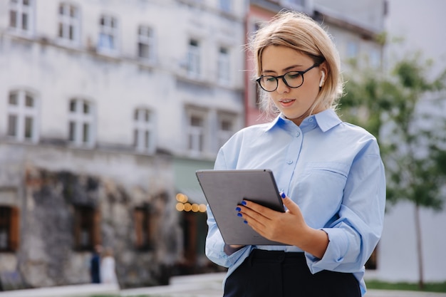 通りに立ってデジタルタブレットを使用して眼鏡とカジュアルウェアの美しい若い女性。都市空間の背景をぼかす。現代の技術コンセプト。