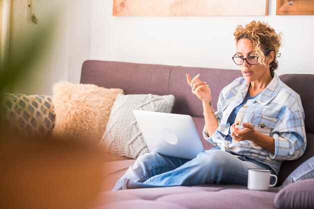 Beautiful young woman in eyeglasses working on laptop while drinking coffee sitting on sofa at home. Businesswoman in casuals and eyeglasses working from home on laptop.