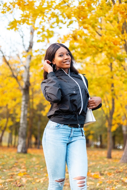 Beautiful young woman in fashionable casual clothes with a jacket, blue jeans and a handbag walks in an autumn park with colored golden foliage