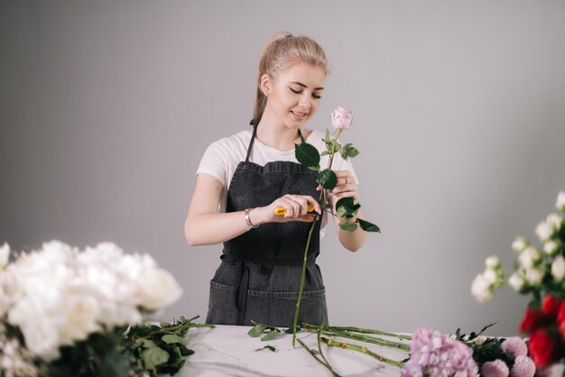 Beautiful young woman florist wearing apron working with fresh rose at the table on white background Concept of working with flowers floral business