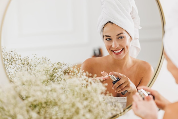 Beautiful young woman in front of mirror applying moisturizer on her face at the beauty salon.