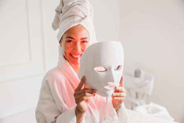 Beautiful young woman getting a led light therapy mask\
treatment for her face at the beauty salon.