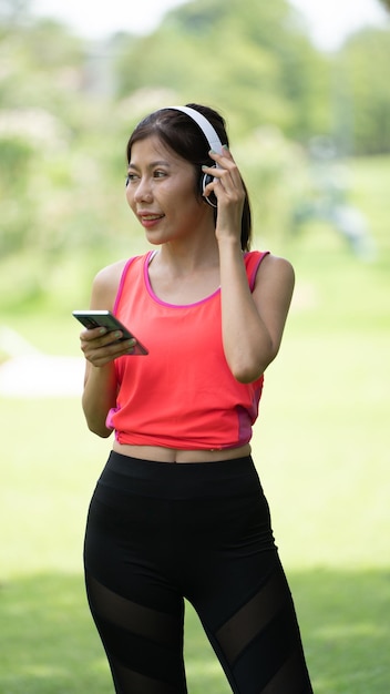 緑の都市公園の背景でポーズをとっているベージュのシャツの美しい若い女性の女の子笑顔の女の子ヘッドフォンで音楽を聴いて幸せに触れる人々のライフスタイルのコンセプトコピースペースをモックアップ