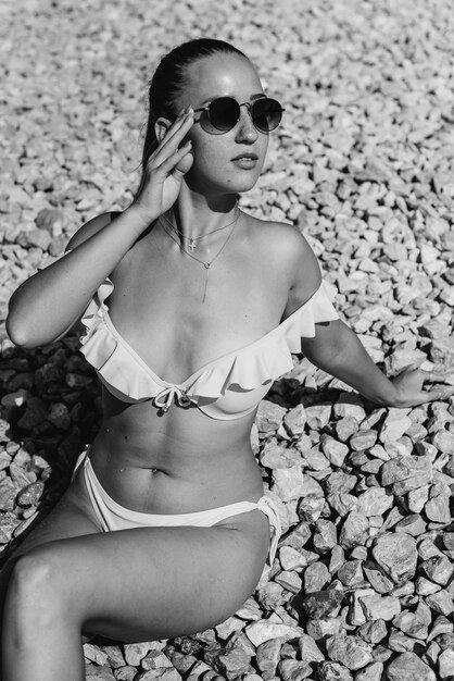 A beautiful young woman in glasses and a bikini is sitting on the ocean shore against the background of huge rocks on a sunny day. Tourism and tourist trips. Black and white.