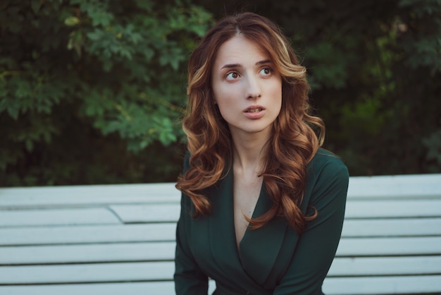 Beautiful young woman in a green dress on a park bench  of bushes