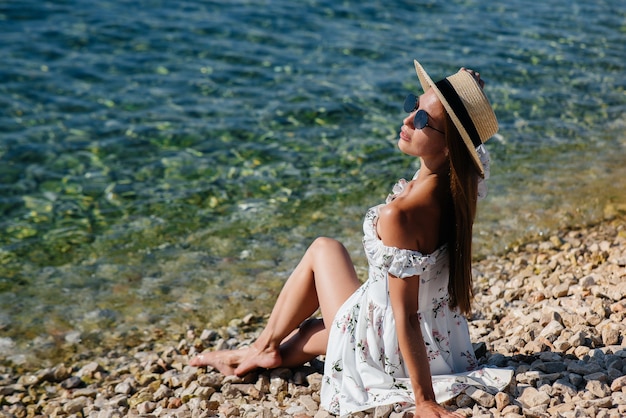 모자, 안경, 가벼운 드레스를 입은 아름다운 젊은 여성이 화창한 날 거대한 바위를 배경으로 바다 해안에 앉아 있습니다. 관광 및 관광 여행.