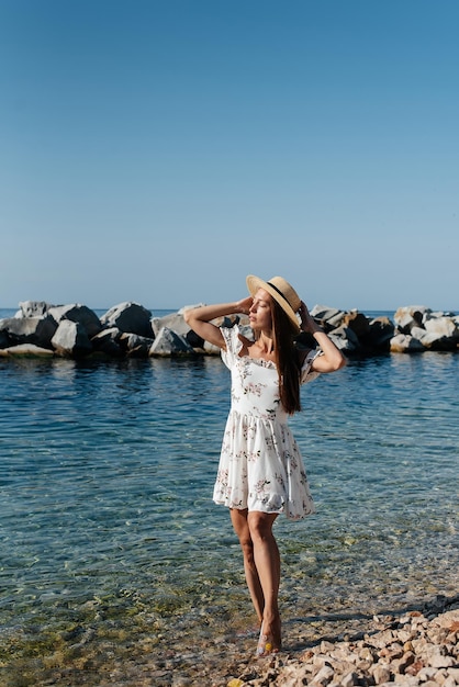 A beautiful young woman in a hat glasses and a light dress is\
walking along the ocean shore against the background of huge rocks\
on a sunny day tourism and tourist trips