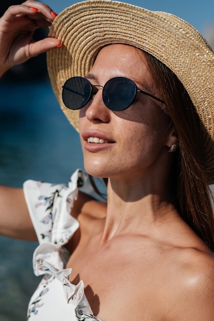 A beautiful young woman in a hat glasses and a light dress is\
walking along the ocean shore against the background of huge rocks\
on a sunny day tourism and tourist trips