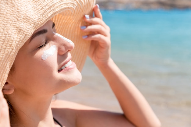 beautiful young woman in hat is applying sunblock under her eyes and on her nose like indian. sun protection concept.