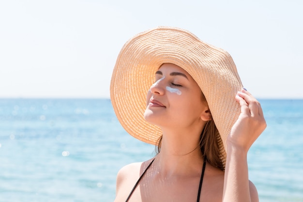 Beautiful young woman in hat is applying sunblock under her eyes and on her nose like Indian. Sun protection concept.