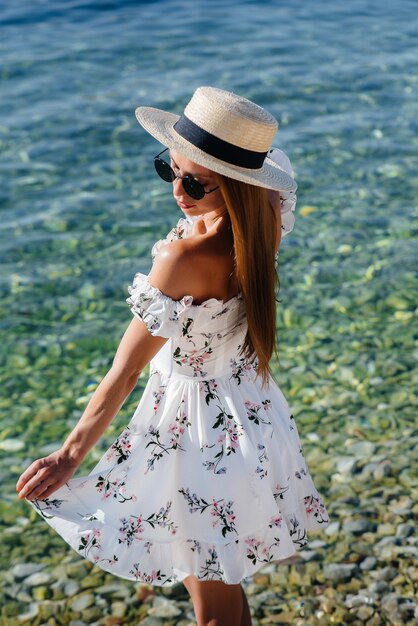 모자를 쓰고 가벼운 드레스를 입은 아름다운 젊은 여성이 화창한 날 거대한 바위를 배경으로 해안을 따라 걷고 있습니다. 관광 및 휴가 여행.