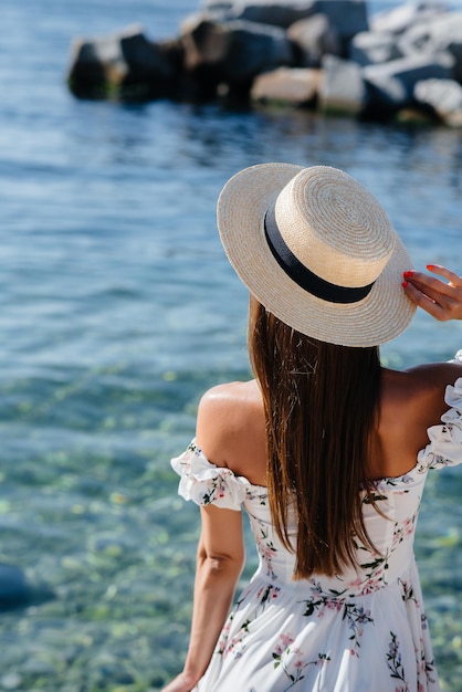 A beautiful young woman in a hat and a light dress with her back is walking along the ocean shore against the background of huge rocks on a sunny day. Tourism and vacation travel.