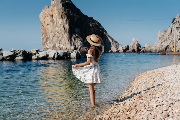 A beautiful young woman in a hat and a light dress with her\
back is walking along the ocean shore against the background of\
huge rocks on a sunny day tourism and vacation travel