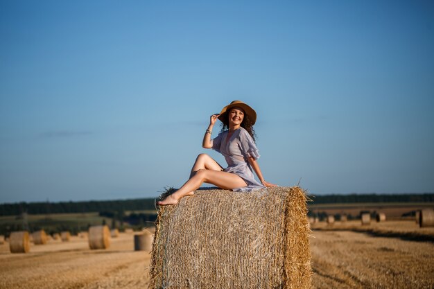 A beautiful young woman in a hat and a summer dress sits on a sheaf of hay in a field. Rural nature, wheat field