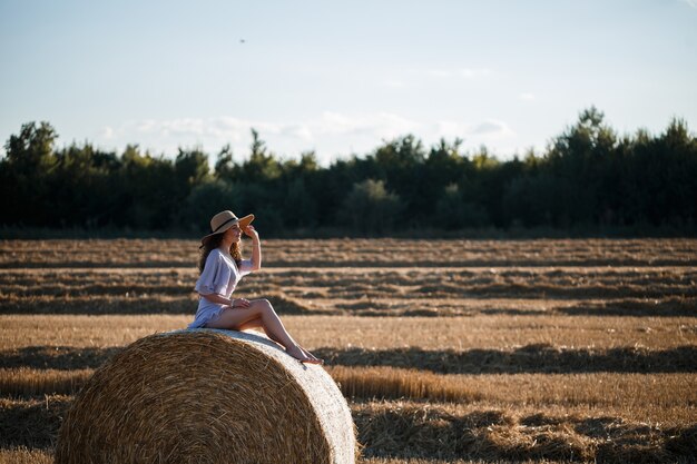 Красивая молодая женщина в шляпе и летнем платье сидит на снопе сена в поле. Сельская природа, пшеничное поле